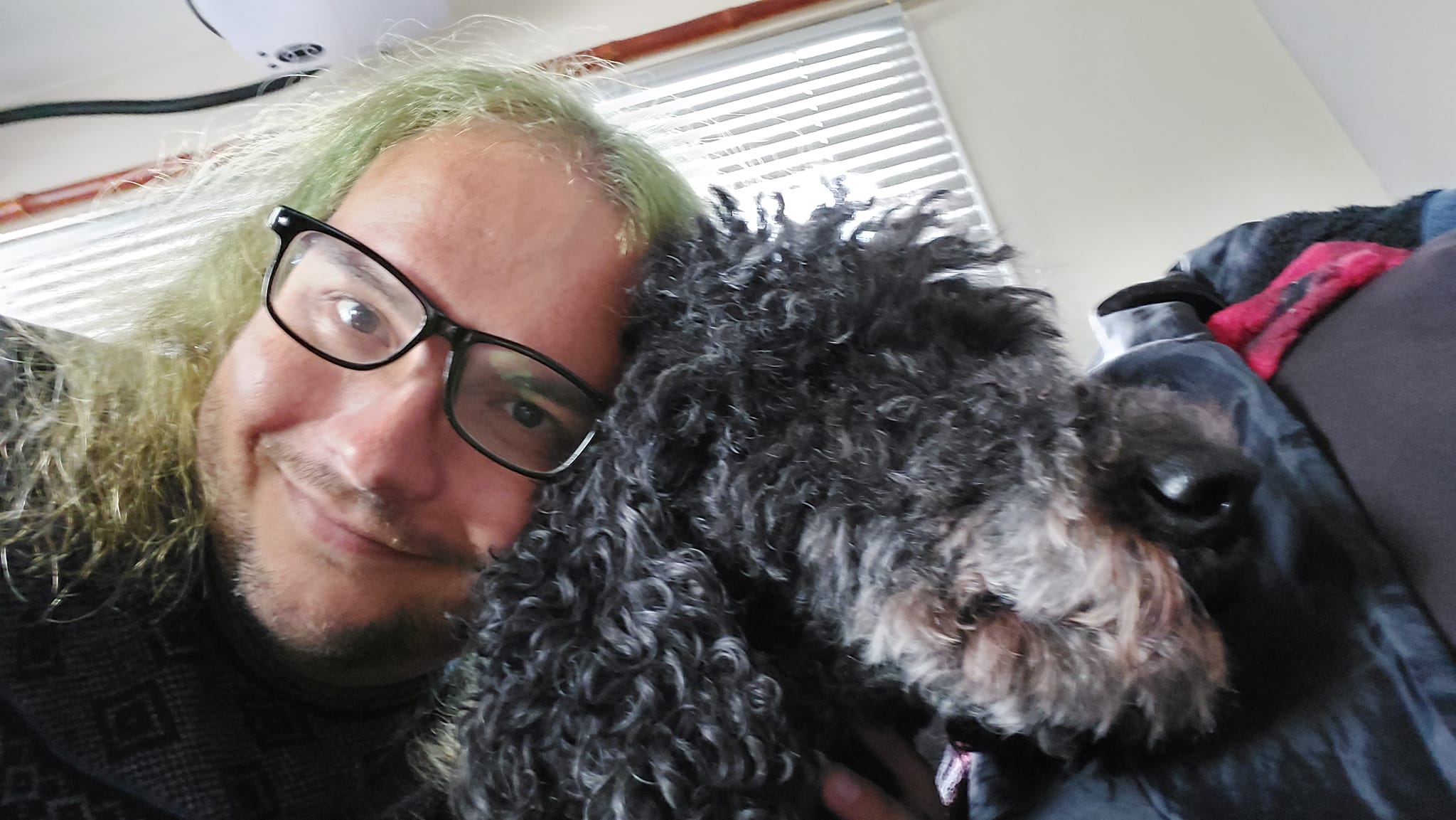 A man with long green hair lays his head next to the head of a black standard poodle, both are comforted